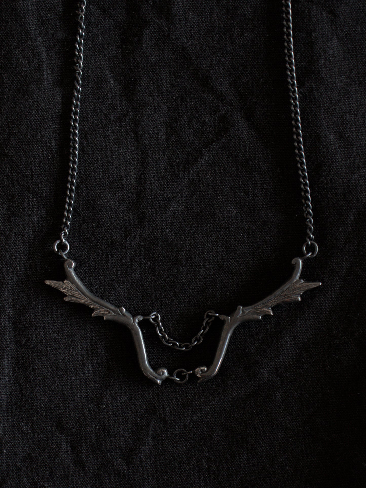 A blackened silver necklace sits on a black fabric background. The necklace has a fine chain with wing shaped pendants joined in the centre with another chain.