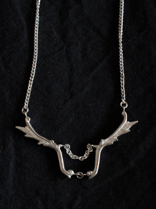 A silver necklace sits on a black fabric background. The necklace has a fine chain with wing shaped pendants joined in the centre with another chain.