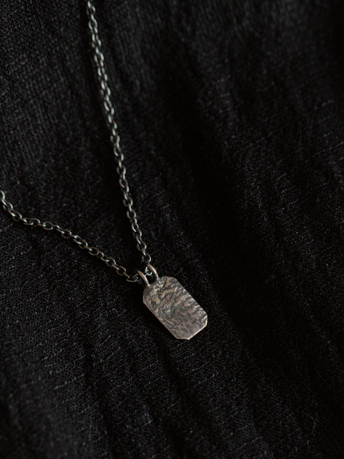 Black fabric background with a blackened silver necklace. The necklace has a fine chain with an octagon shaped pendant with textured surface.
