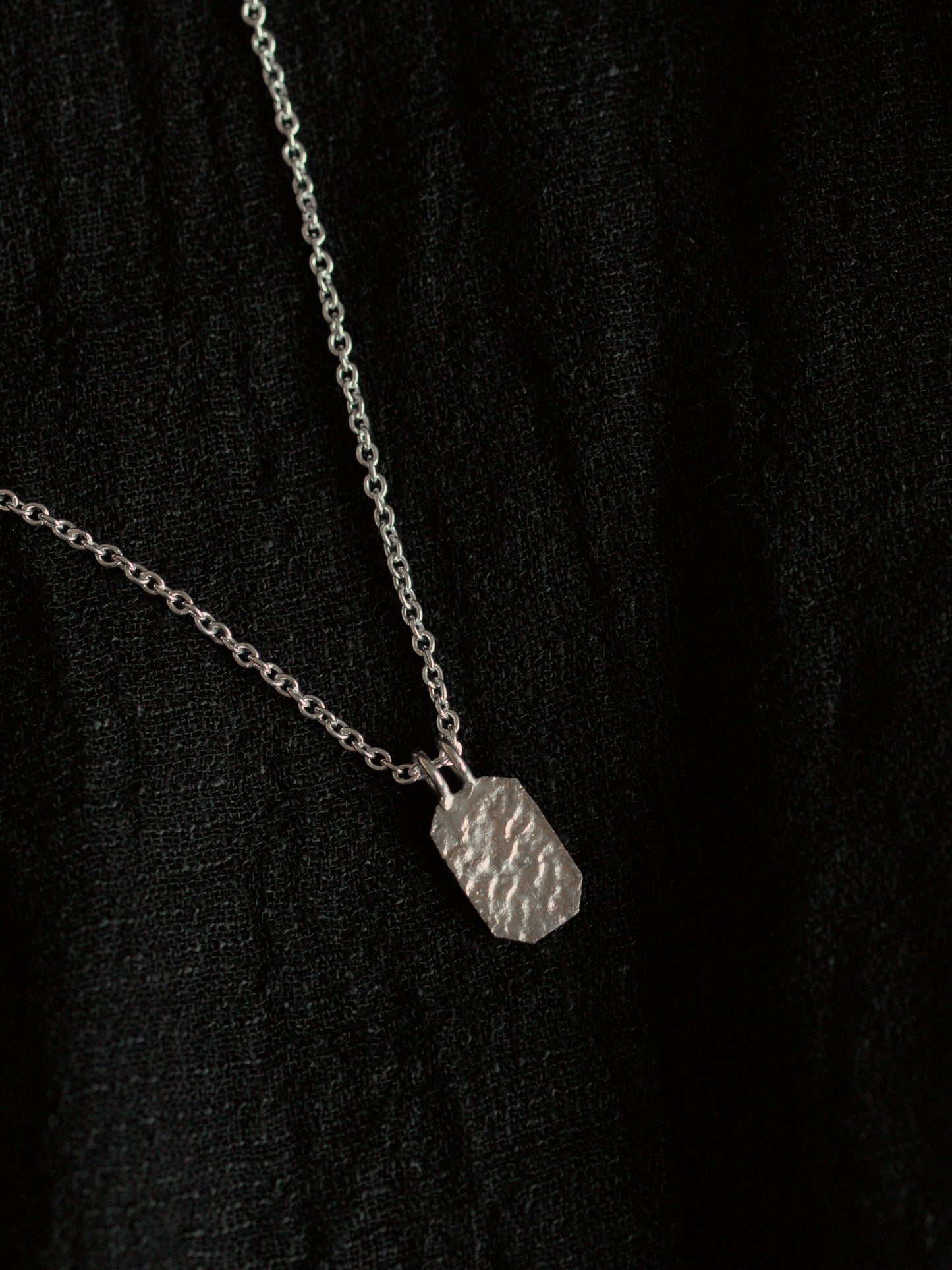 Black fabric background with a silver necklace. The necklace has a fine chain with an octagon shaped pendant with textured surface.