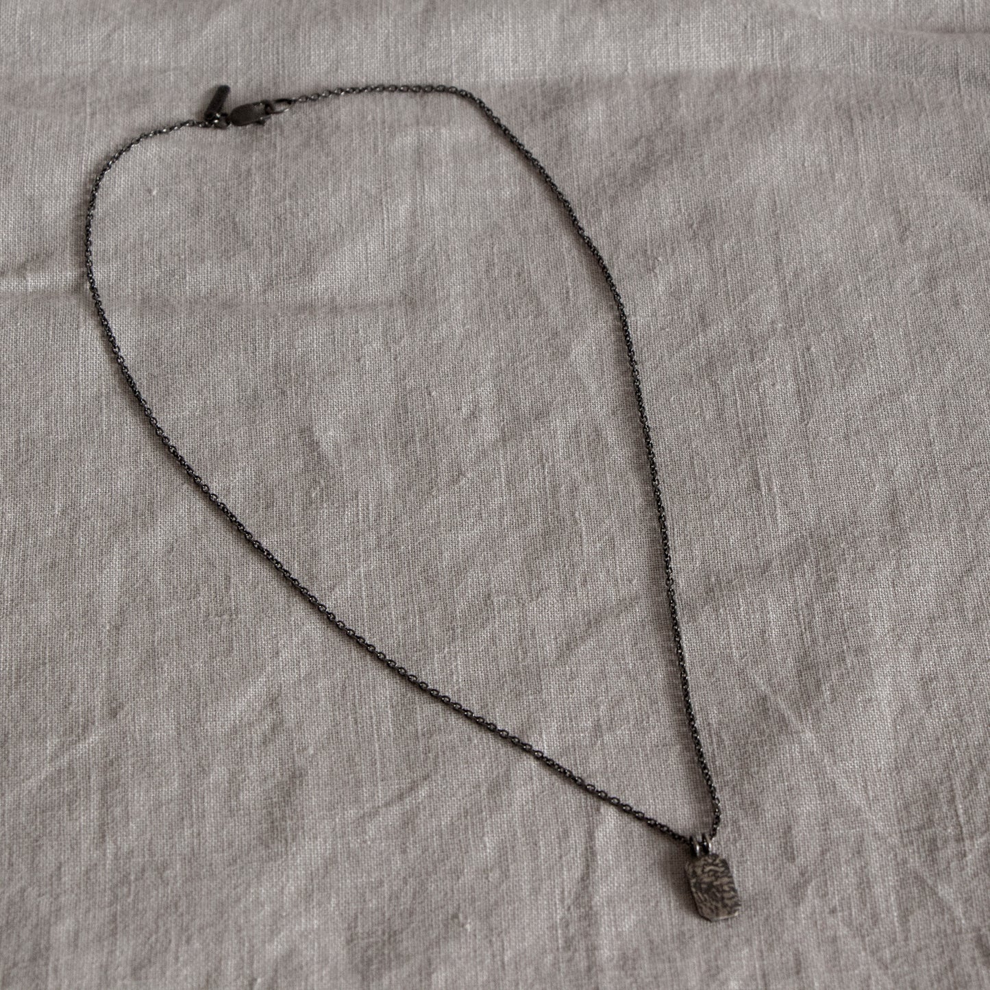 Grey fabric background with a blackened silver necklace shown in full. The necklace has a fine chain and clasp on one end and an octagon shaped pendant with textured surface on the other end.