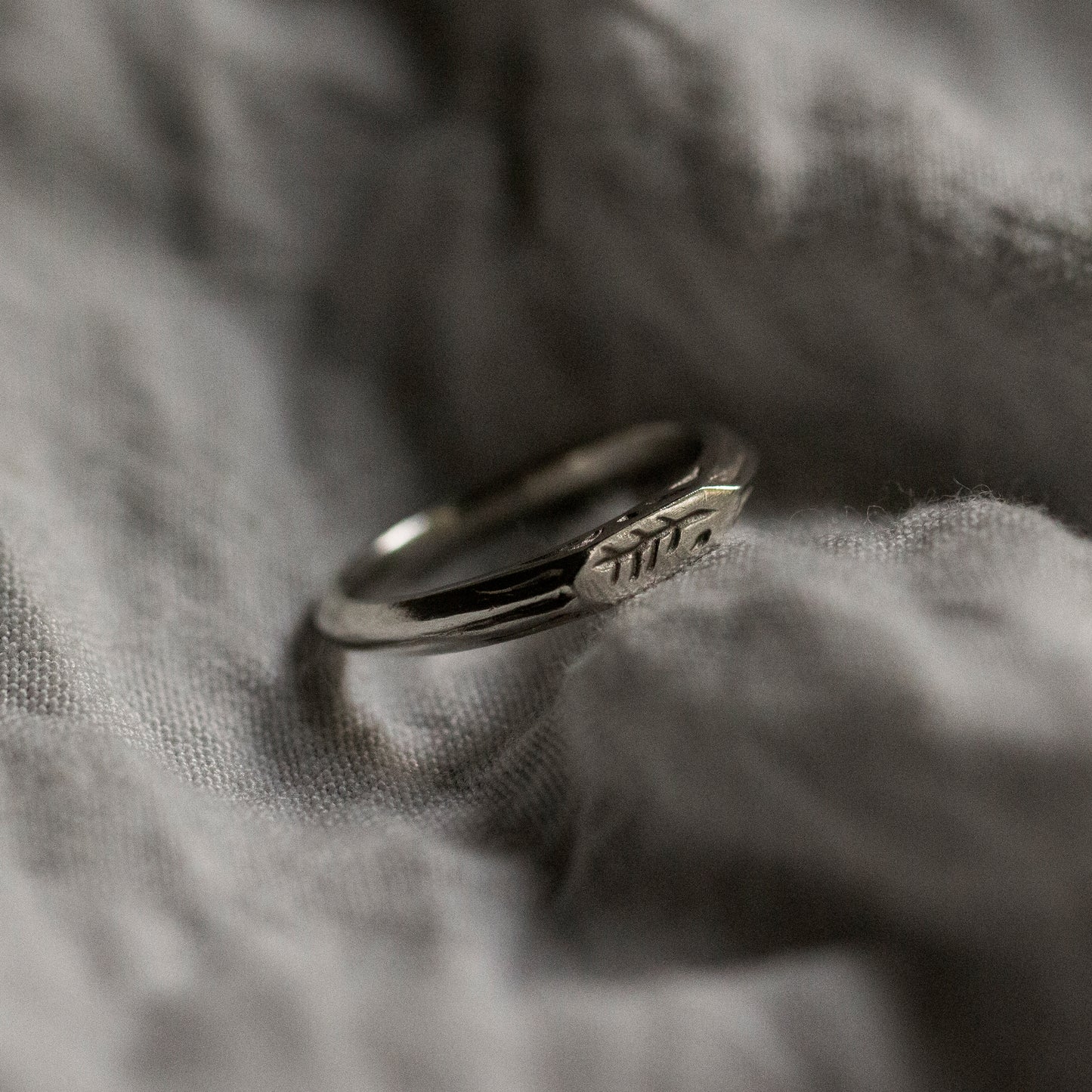 Grey fabric background with a silver ring in the centre. The ring is narrow in width and has a geometric shape with several embellishments engraved into the sides.