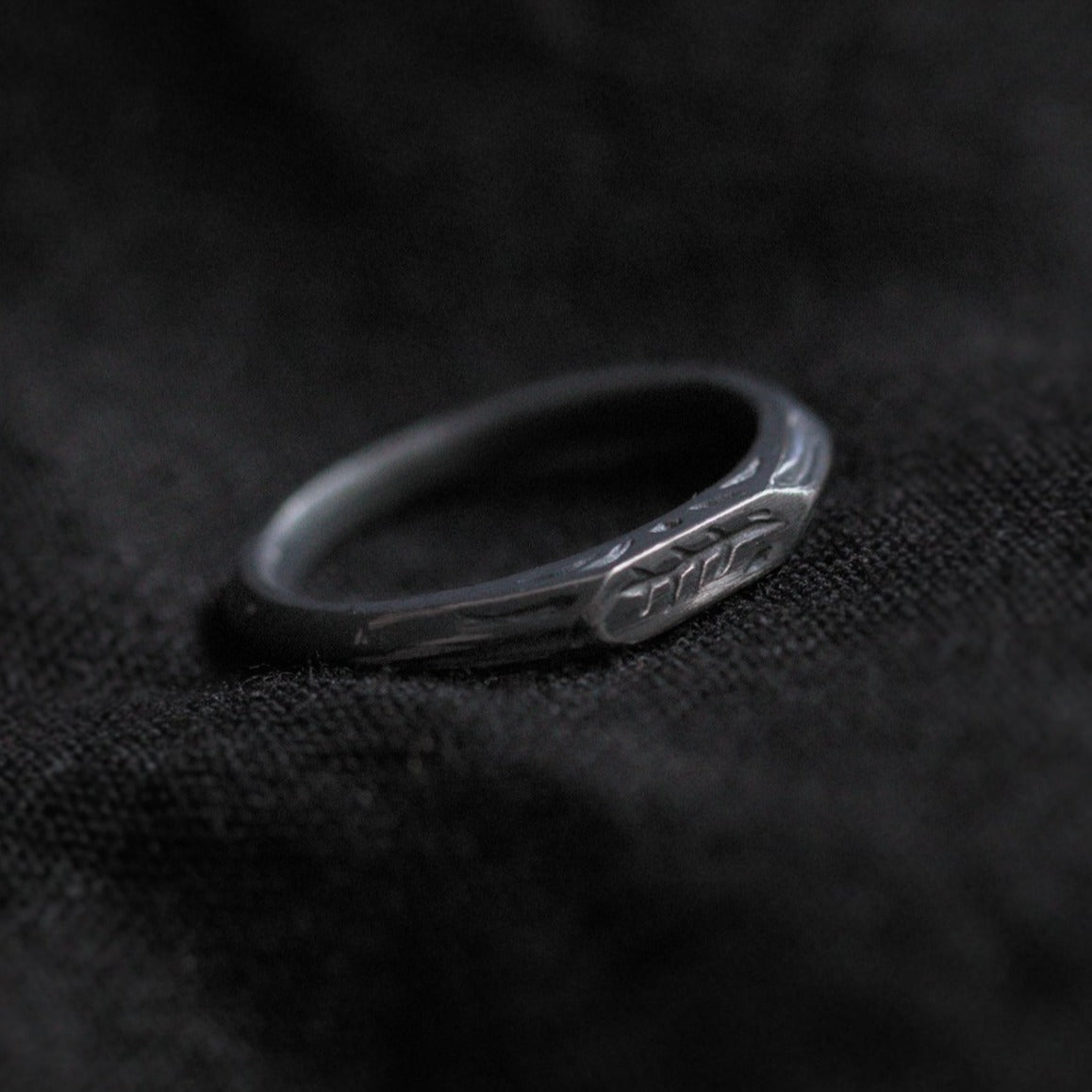 Black fabric background with a blackened silver ring in the centre. The ring is narrow in width and has a geometric shape with several embellishments engraved into the sides. The top of the ring is most visible and shows a twig engraving in the centre.
