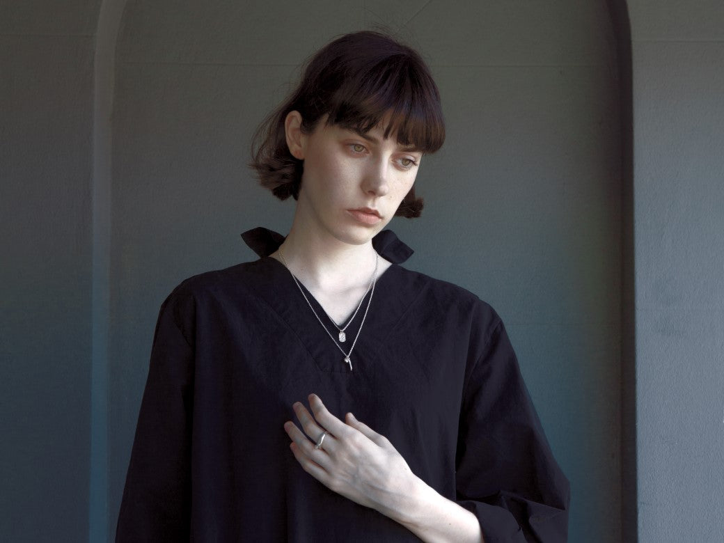 Stylised image of a model with brown short hair wearing a black shirt and two silver necklaces. The model's hand is on her chest, showing a silver ring on one finger. The background is a dark grey wall with part of an arch detail on the wall.
