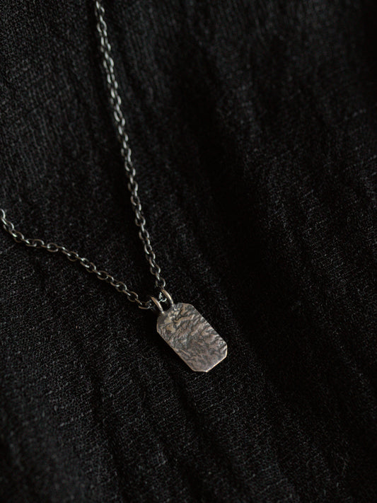 Black fabric background with a blackened silver necklace. The necklace has a fine chain with an octagon shaped pendant with textured surface.