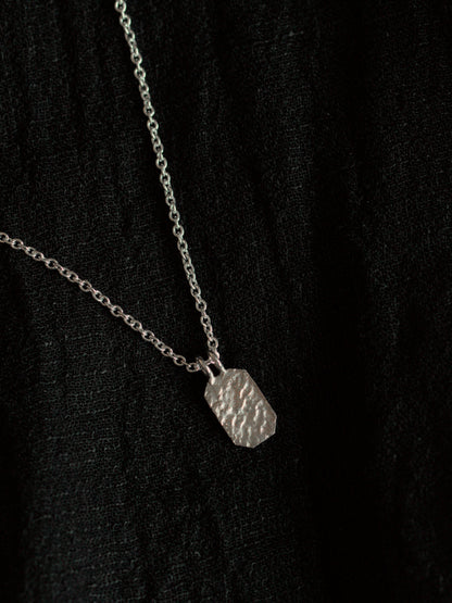 Black fabric background with a silver necklace. The necklace has a fine chain with an octagon shaped pendant with textured surface.
