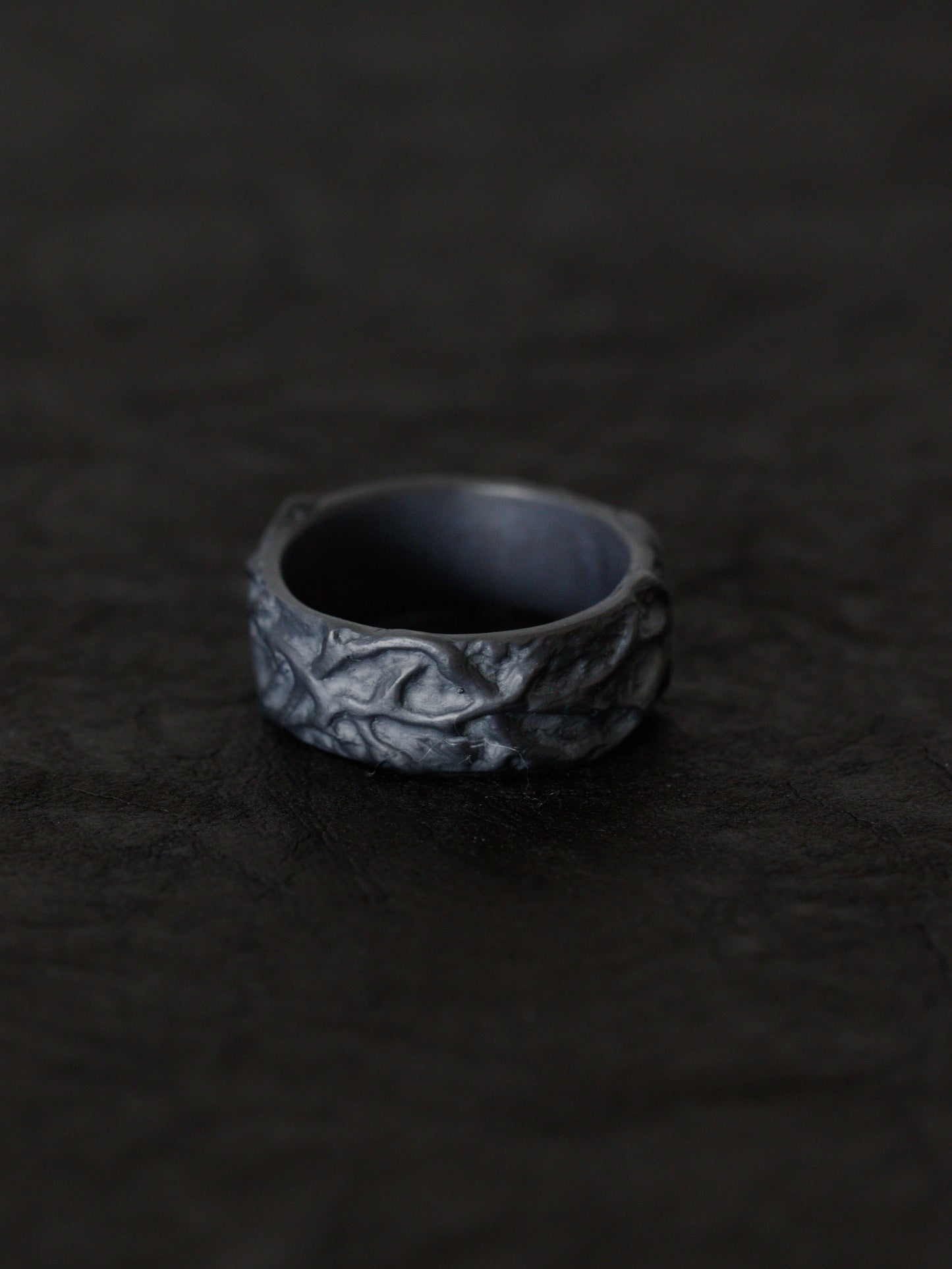 A blackened silver ring with an organic, textured, thorn inspired pattern, resting on a dark, textured surface.