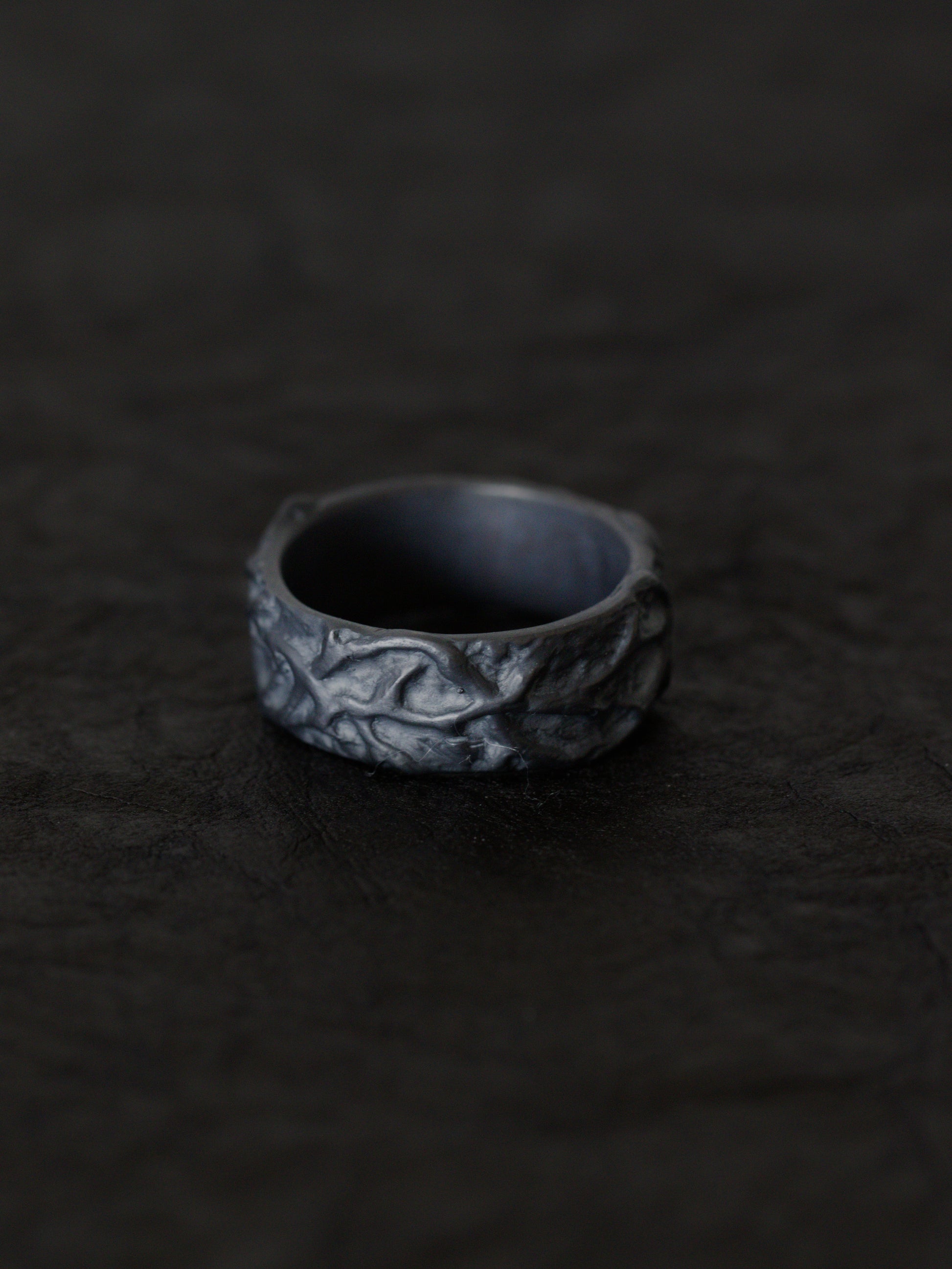 A blackened silver ring with an organic, textured, thorn inspired pattern, resting on a dark, textured surface.