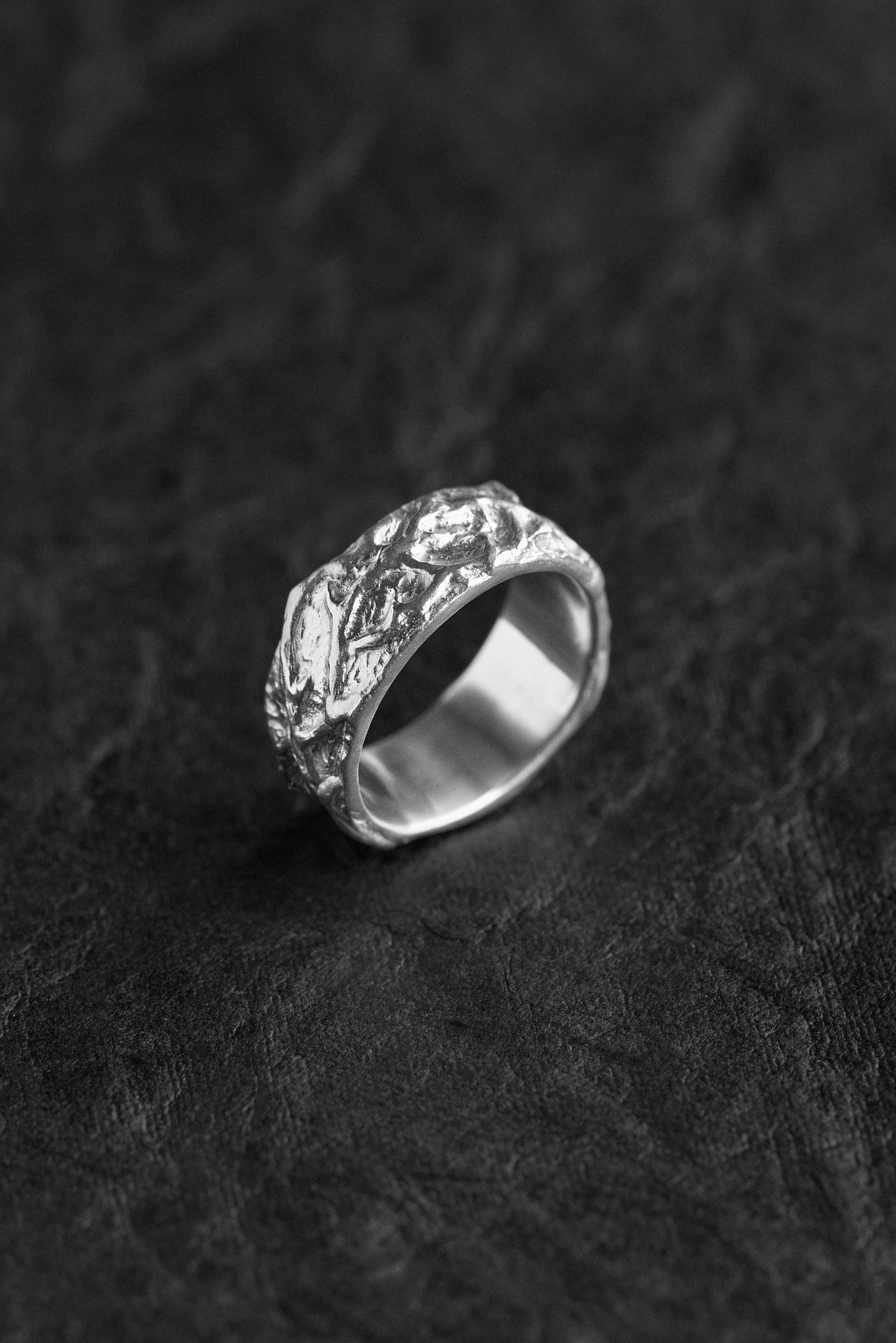 A silver ring with an organic, textured, tree-bark-like pattern, resting on a dark textured surface.