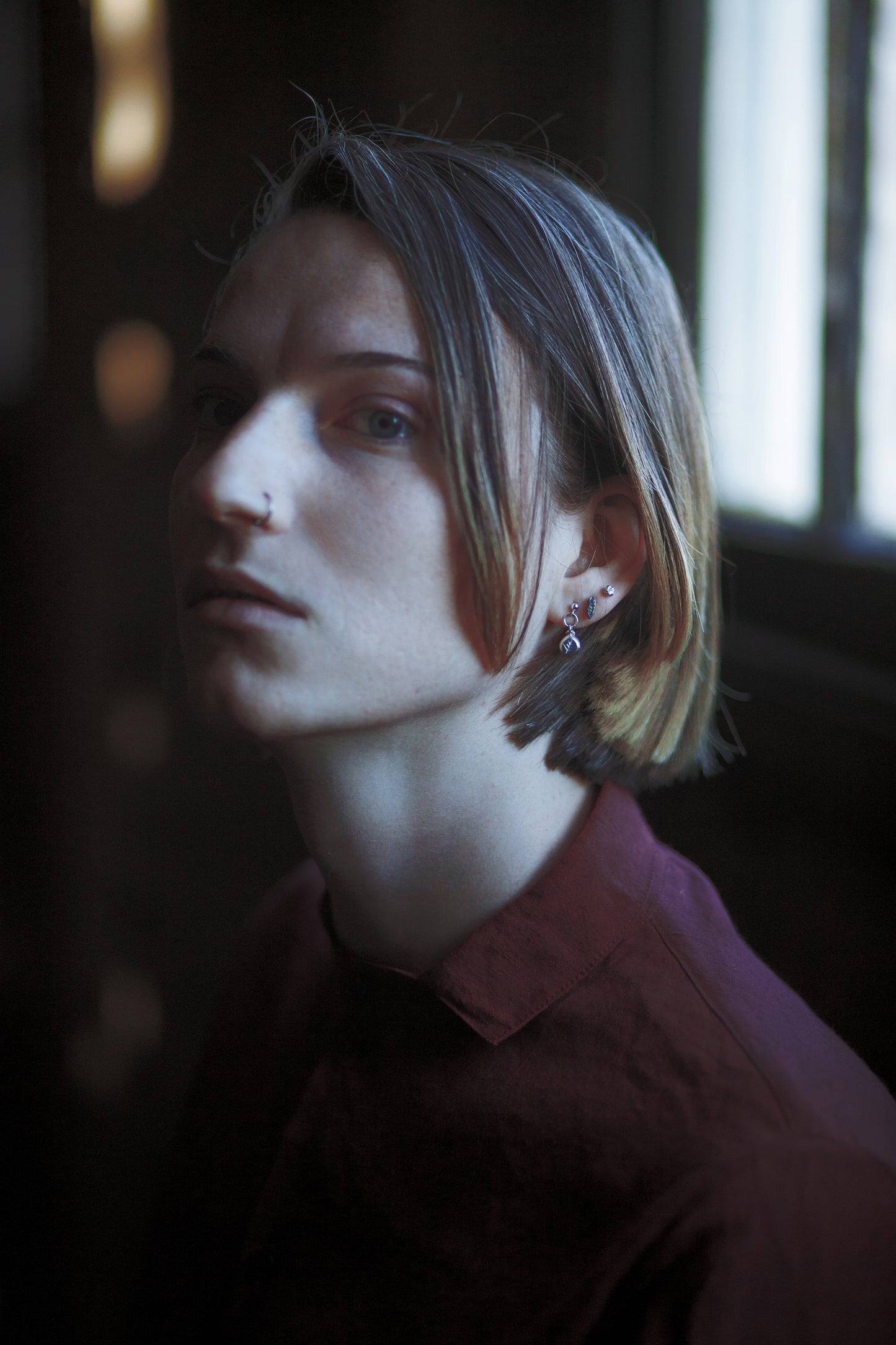 The model stands in the mid ground, with an indoor setting in the background. The model has a short haircut and is wearing three pairs of earrings on her ear and a dark red, collared shirt.