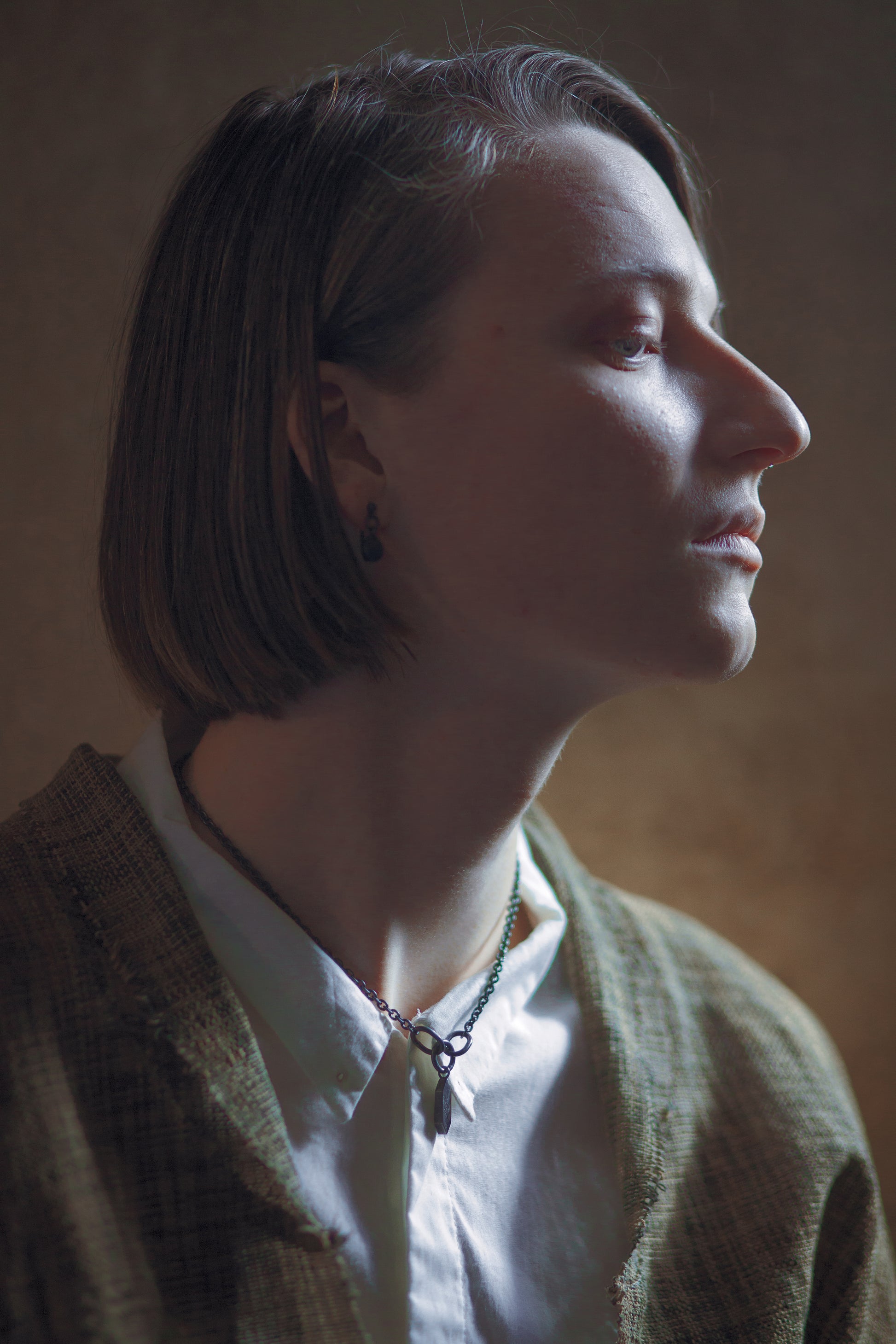 A stylised image of a model wearing a white shirt and brown jacket standing on a brown backgroun. The model is also wearing a blackened silver earring and blackened silver necklace.