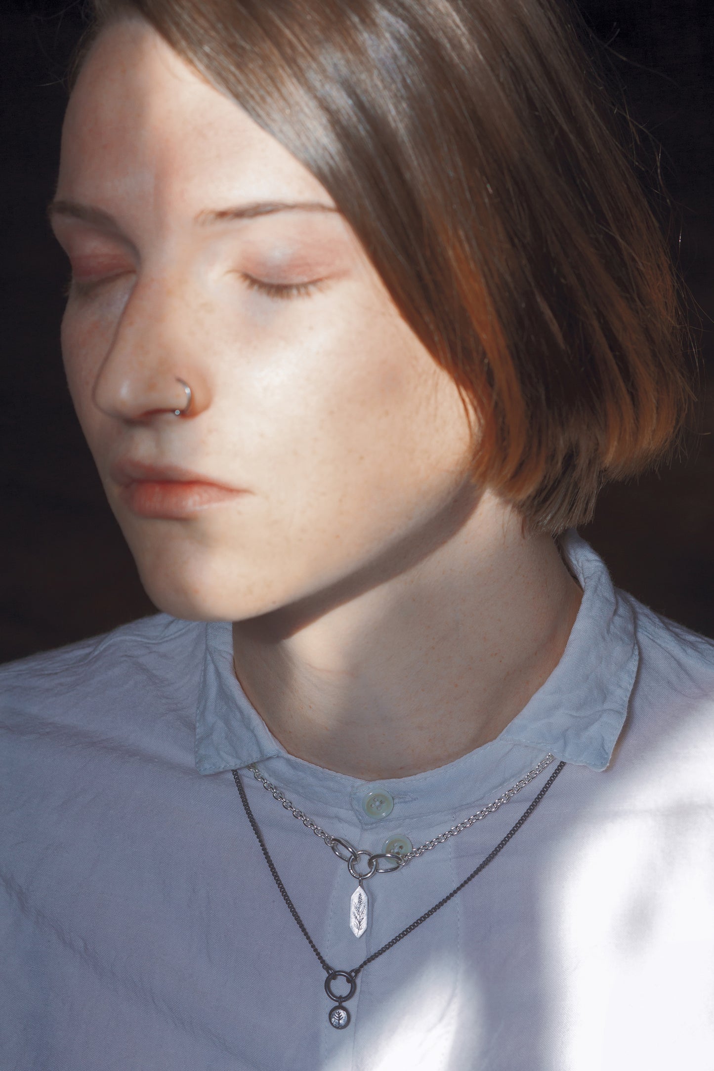 A stylised image of a model wearing two necklaces. one necklace is a bright silver tone with a geometric pendant, and the necklace below it is a blackened silver tone with a round pendant. The model has her eyes closed and is wearing a white button up shirt with a collar. Parts of the necklaces are covered by the shirt's collar.