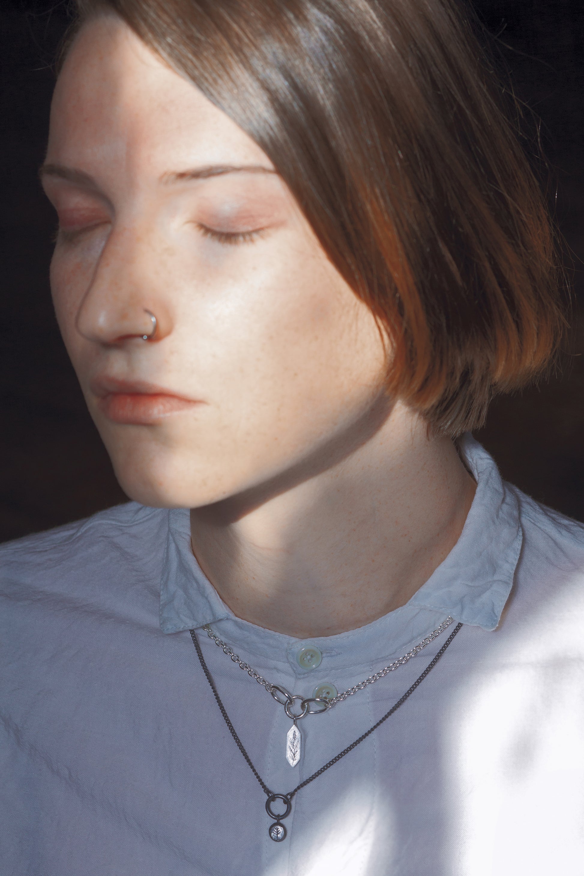 A stylised image of a model wearing two necklaces. one necklace is a bright silver tone with a geometric pendant, and the necklace below it is a blackened silver tone with a round pendant. The model has her eyes closed and is wearing a white button up shirt with a collar. Parts of the necklaces are covered by the shirt's collar.