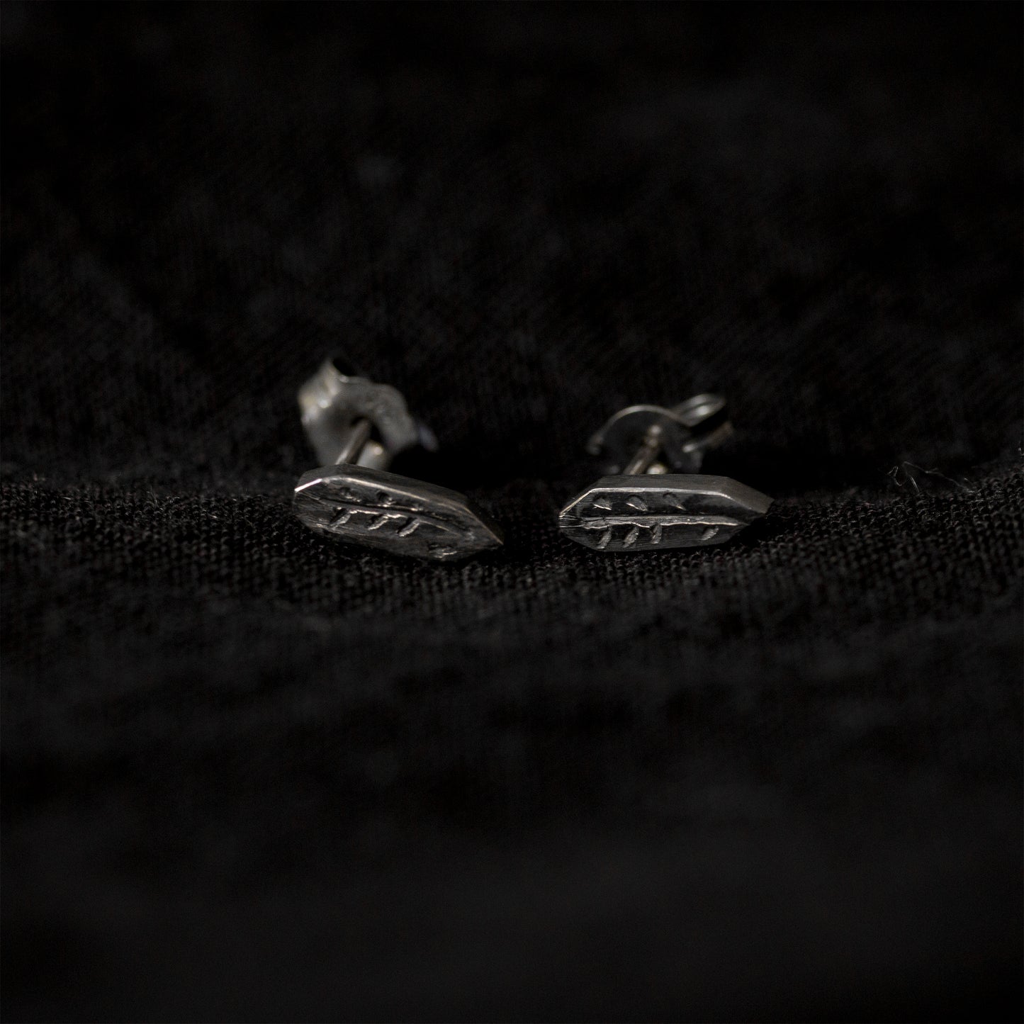 Black fabric background with a pair of blackened silver stud earrings lying in the middle of the image. The earrings are an irregular geometric shape with a carving of a twig in the middle.