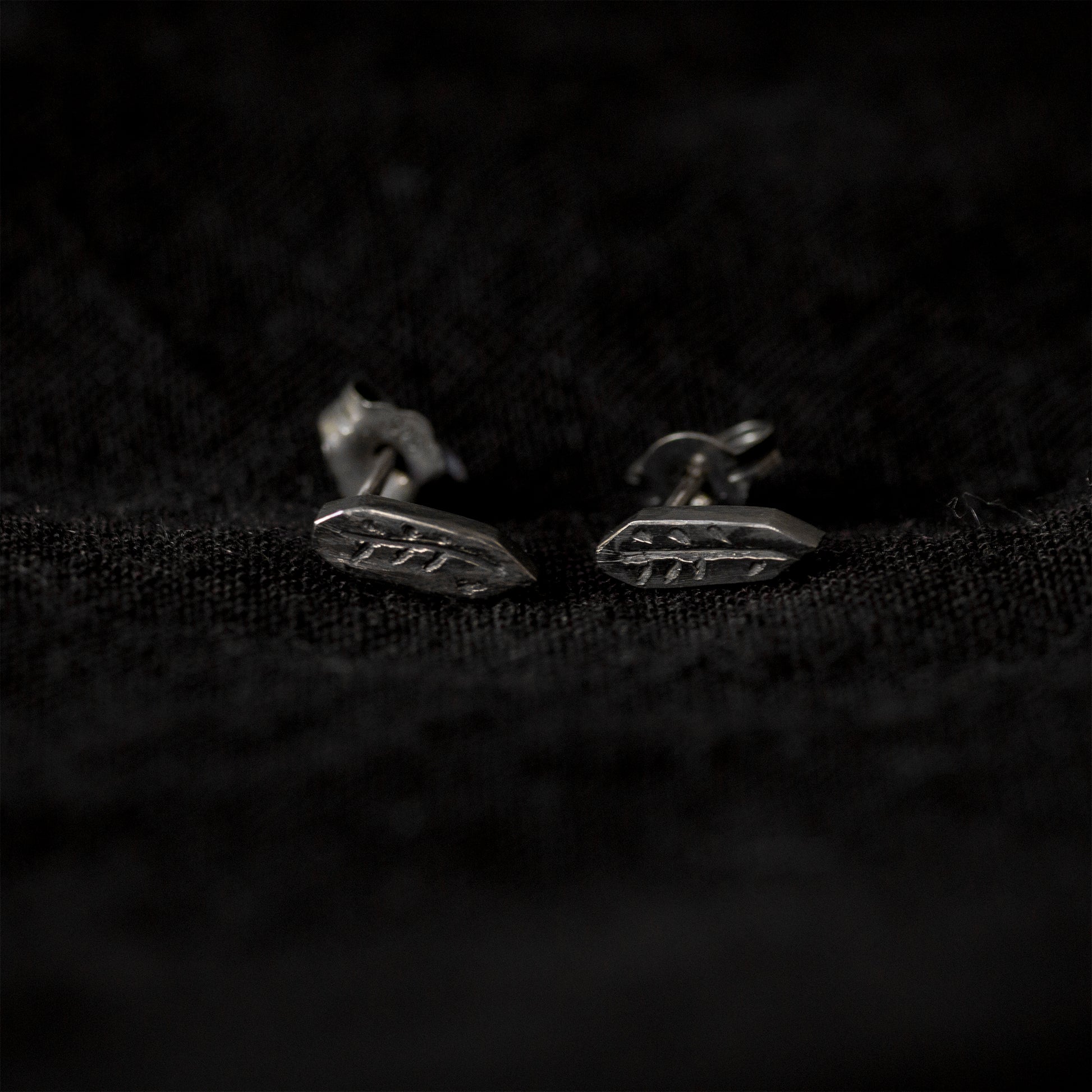 Black fabric background with a pair of blackened silver stud earrings lying in the middle of the image. The earrings are an irregular geometric shape with a carving of a twig in the middle.