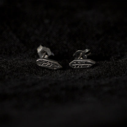 Black fabric background with a pair of blackened silver stud earrings lying in the middle of the image. The earrings are an irregular geometric shape with a carving of a twig in the middle.