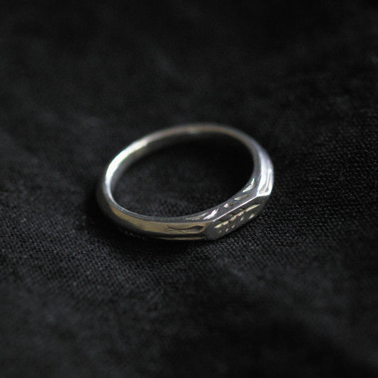 Black fabric background with a silver ring in the centre. The ring is narrow in width and has a geometric shape with several embellishments engraved into the sides.