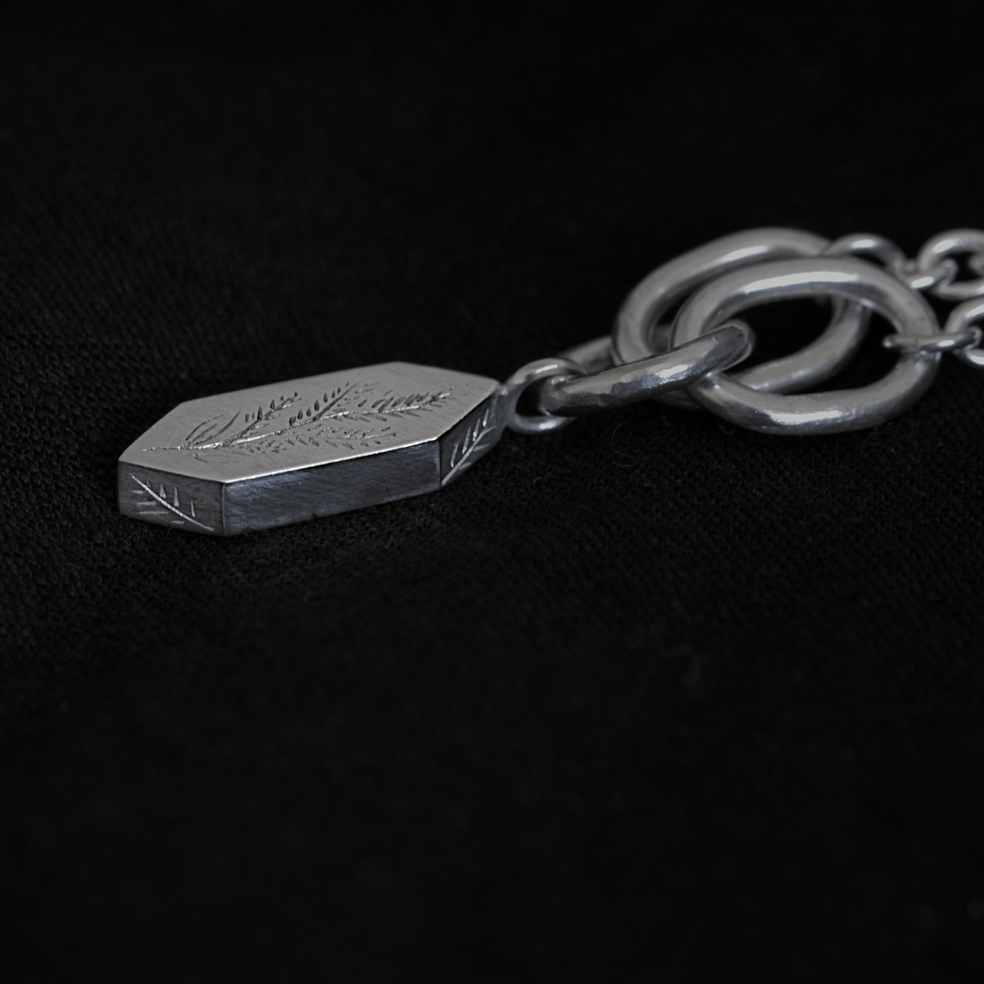 A close up and side view of the silver necklace and the pendant, which sits on a black fabric background. The image shows parts of the necklace's chain, with three larger rings that connect the geometric pendant. The penant features engravings of a plant and some leaves on the side.