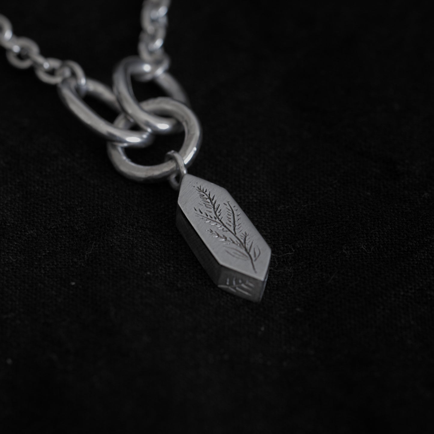 A close up of the silver necklace and the pendant sit on a black fabric background. The image shows parts of the necklace's chain, with three larger rings that connect the geometric pendant. The penant features engravings of a plant and some leaves on the side.