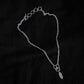 Black fabric background with a silver necklace in the centre. The necklace features five rings and a clasp, the chain has a narrow width, and the chains meet at three larger rings that connect a geometric shaped pendant with an engraving of a plant on the surface.