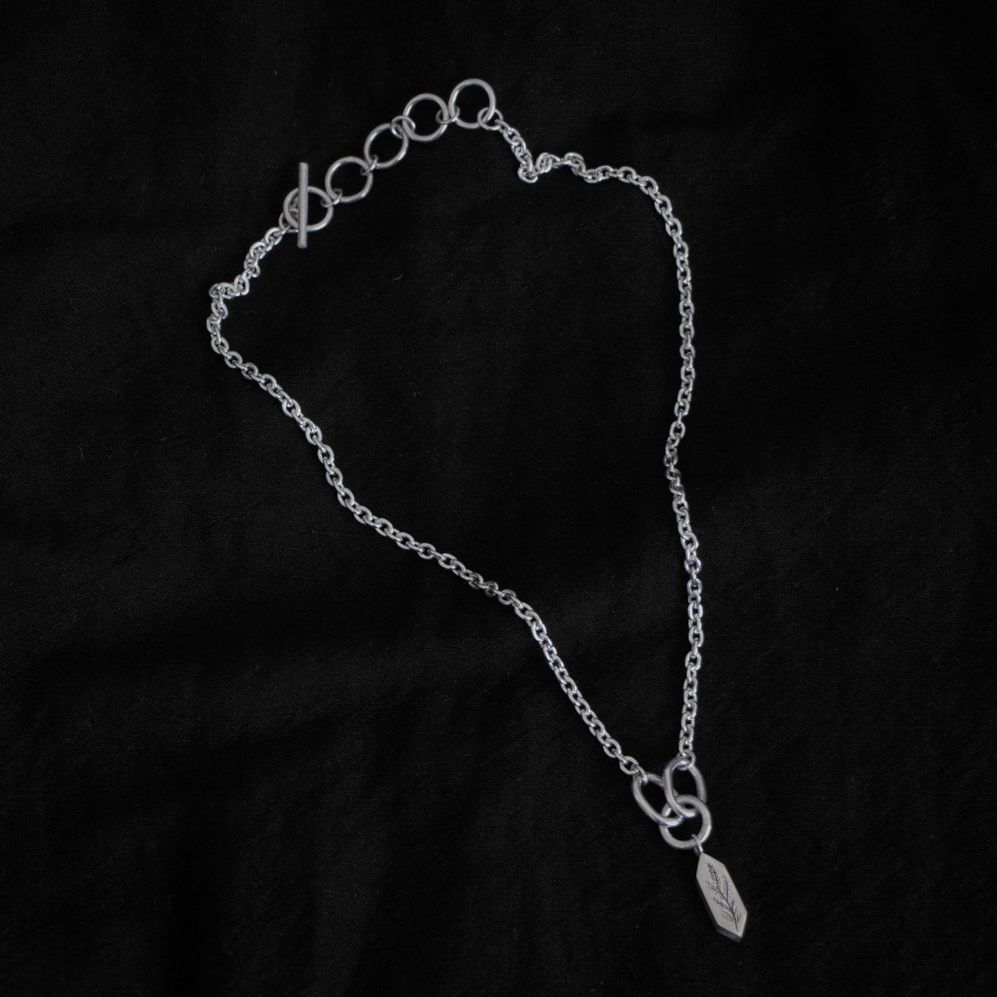 Black fabric background with a silver necklace in the centre. The necklace features five rings and a clasp, the chain has a narrow width, and the chains meet at three larger rings that connect a geometric shaped pendant with an engraving of a plant on the surface.