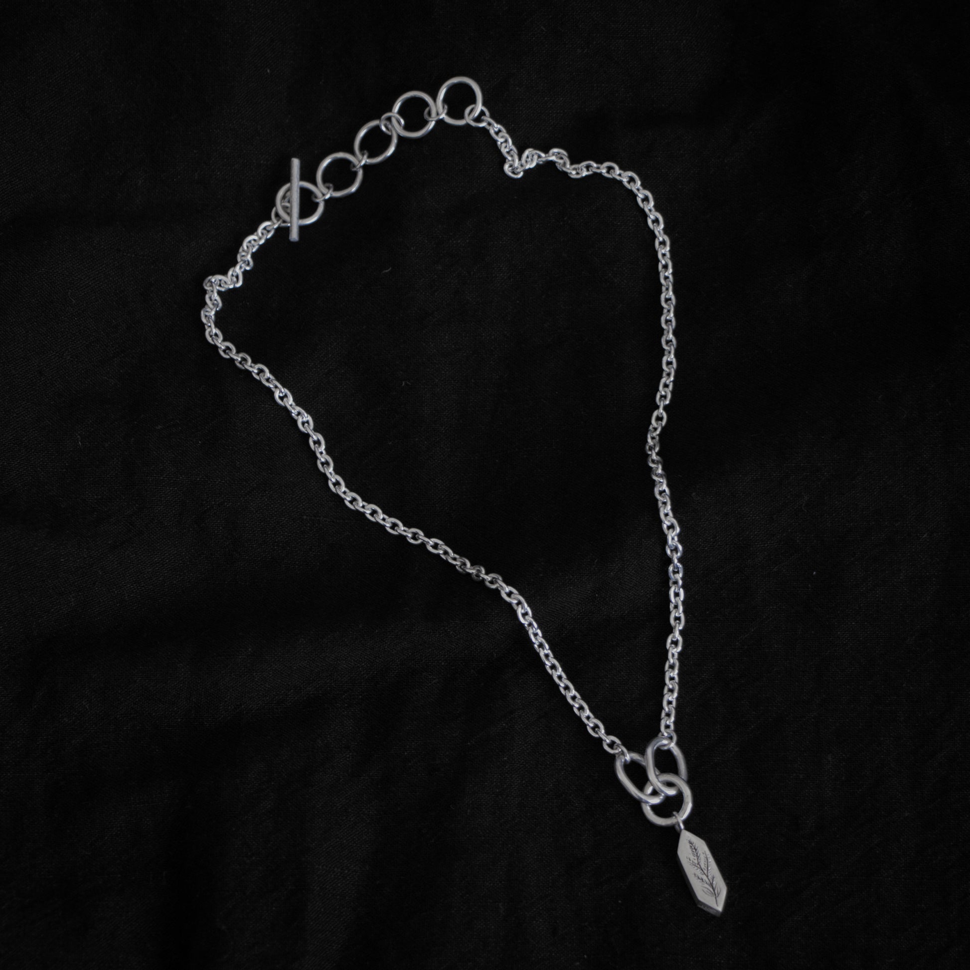 Black fabric background with a silver necklace in the centre. The necklace features five rings and a clasp, the chain has a narrow width, and the chains meet at three larger rings that connect a geometric shaped pendant with an engraving of a plant on the surface.