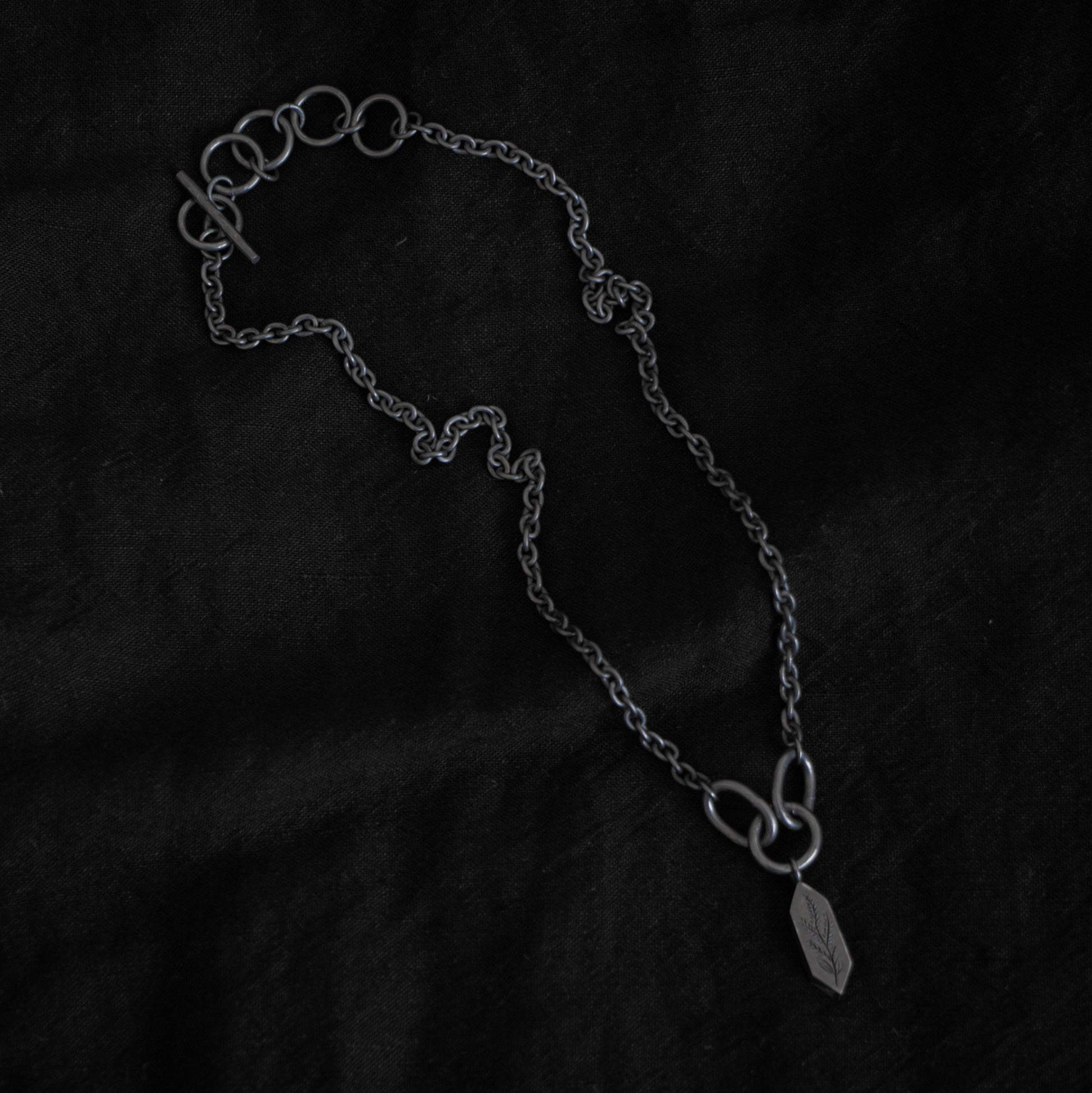 Black fabric background with a blackened silver necklace in the centre. The necklace features five rings and a clasp, the chain has a narrow width, and the chains meet at three larger rings that connect a geometric shaped pendant with an engraving of a plant on the surface.
